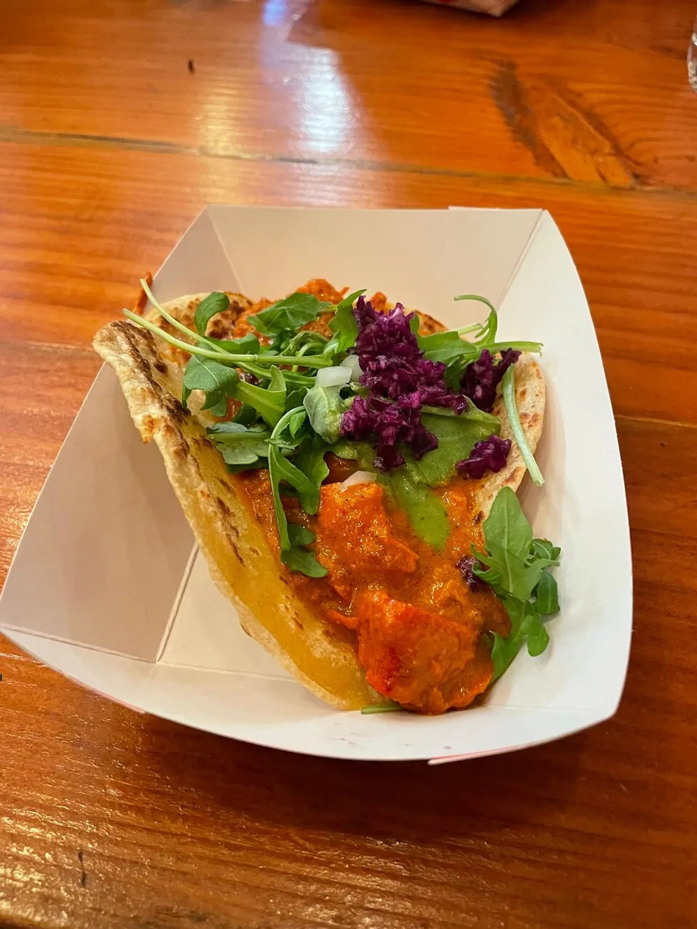 The image shows a colorful taco with a variety of fillings including what appears to be a creamy sauce and fresh greens served in a white paper tray on a wooden table