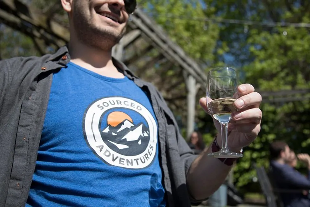 A person is holding a glass of white wine partially cropped from the frame with a joyful expression in an outdoor setting featuring the logo SOURCED ADVENTURES on their T-shirt