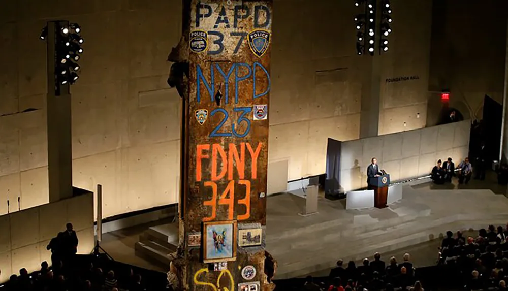 The image shows an interior view of a solemn event with a speaker at a podium and a rusted damaged column with numbers and patches commemorating NYPD FDNY and PAPD members lost likely a relic from the 911 attacks exhibited to honor their sacrifice