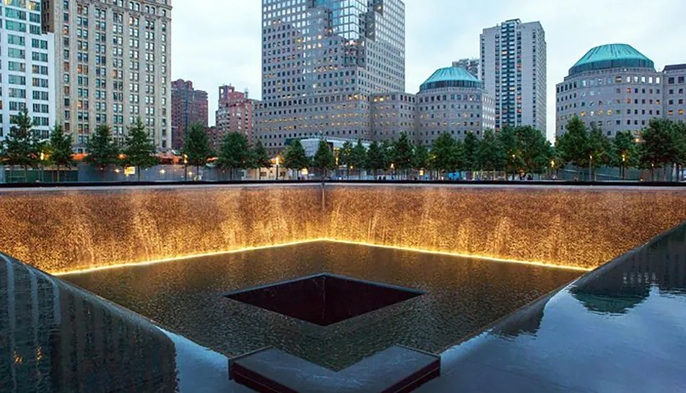 The image shows one of the 911 Memorial pools at twilight surrounded by trees with skyscrapers in the background creating a serene reflection space in memory of the victims of September 11 2001