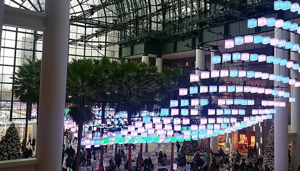 The image shows an indoor space with a large multi-colored cube-shaped light installation suspended from the ceiling surrounded by palm trees and a crowd of people with a festive atmosphere suggestive of a public venue like a mall or event space