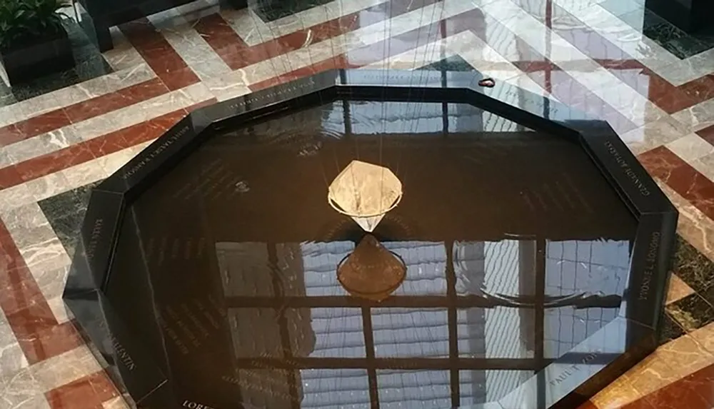 The image shows an octagonal water feature with a glass sculpture in the center reflecting the ceiling and surrounding architecture