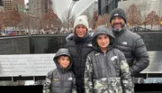 A family of four, dressed in winter clothing, is smiling for the photo in front of the 9/11 Memorial reflecting pools in New York City.