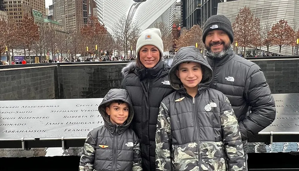 A family of four dressed in winter clothing is smiling for the photo in front of the 911 Memorial reflecting pools in New York City