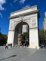 The image shows the Washington Square Arch in New York City on a sunny day with people walking around the open plaza.