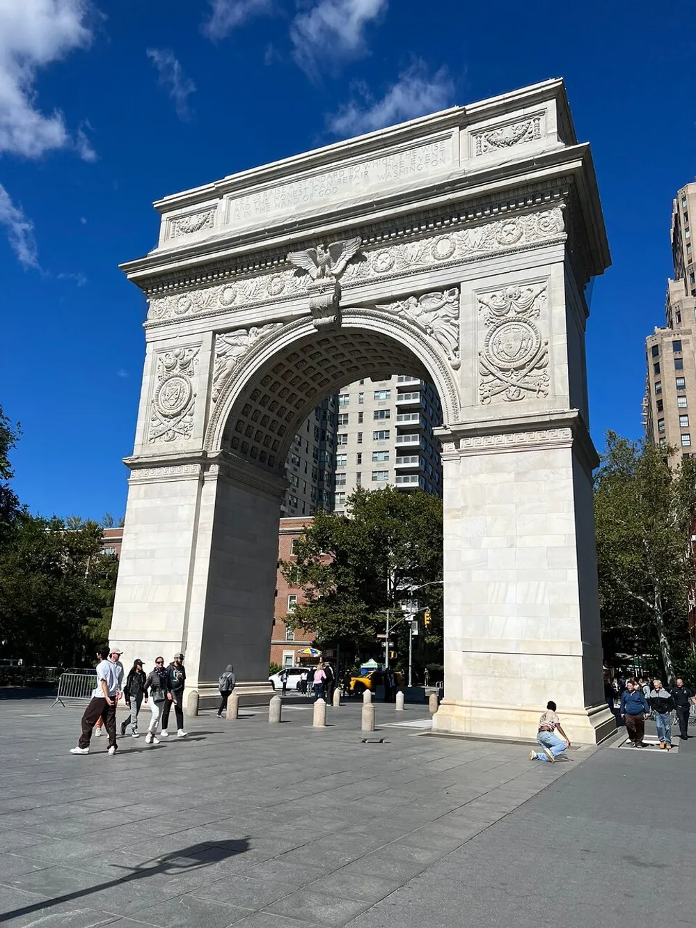 The image shows the Washington Square Arch in New York City on a sunny day with people walking around the open plaza