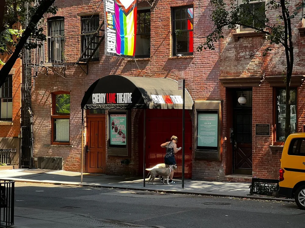 A person and a dog stand outside the Cherry Lane Theatre which displays a rainbow flag on a sunny day with dappled shade from trees