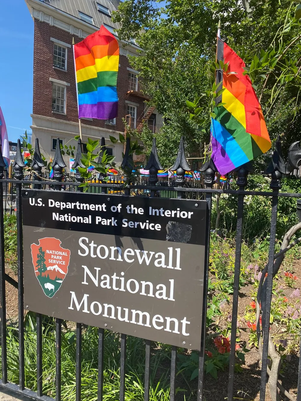 The image shows a sign for the Stonewall National Monument part of the US Department of the Interior National Park Service adorned with multiple rainbow pride flags fluttering in the foreground