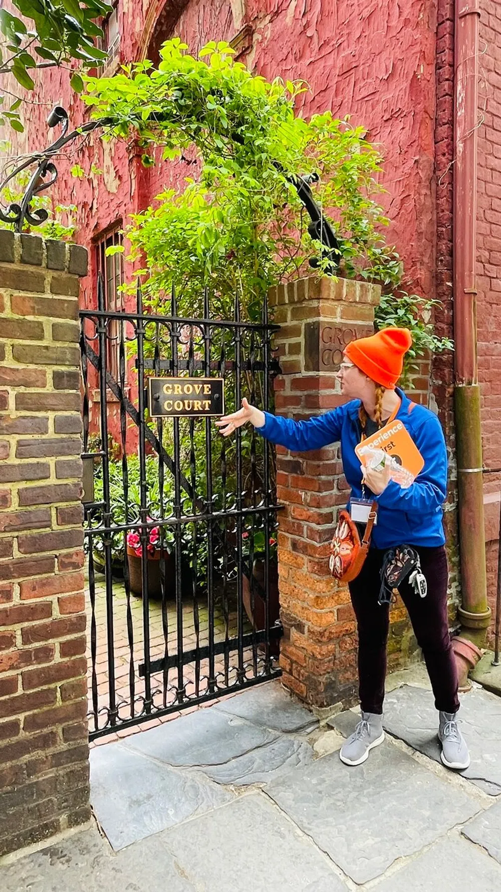 A person in a bright orange beanie is pointing at the Grove Court sign on a black iron gate in front of a charming brick walled alcove with green plants