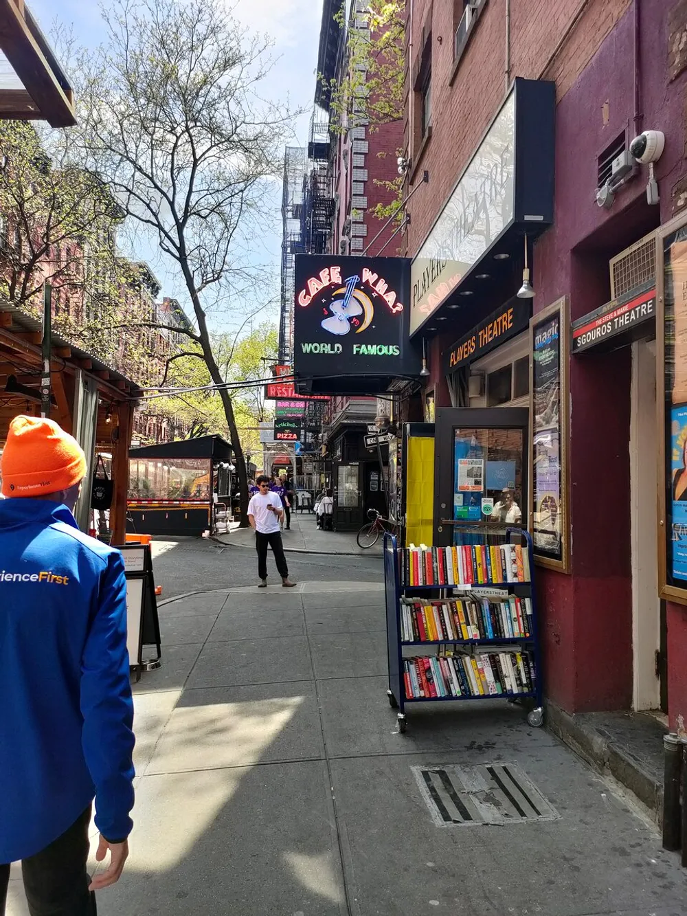 The image shows a bustling street scene with various signs including one for Caf Wha claiming to be world-famous and people walking by a stand filled with books