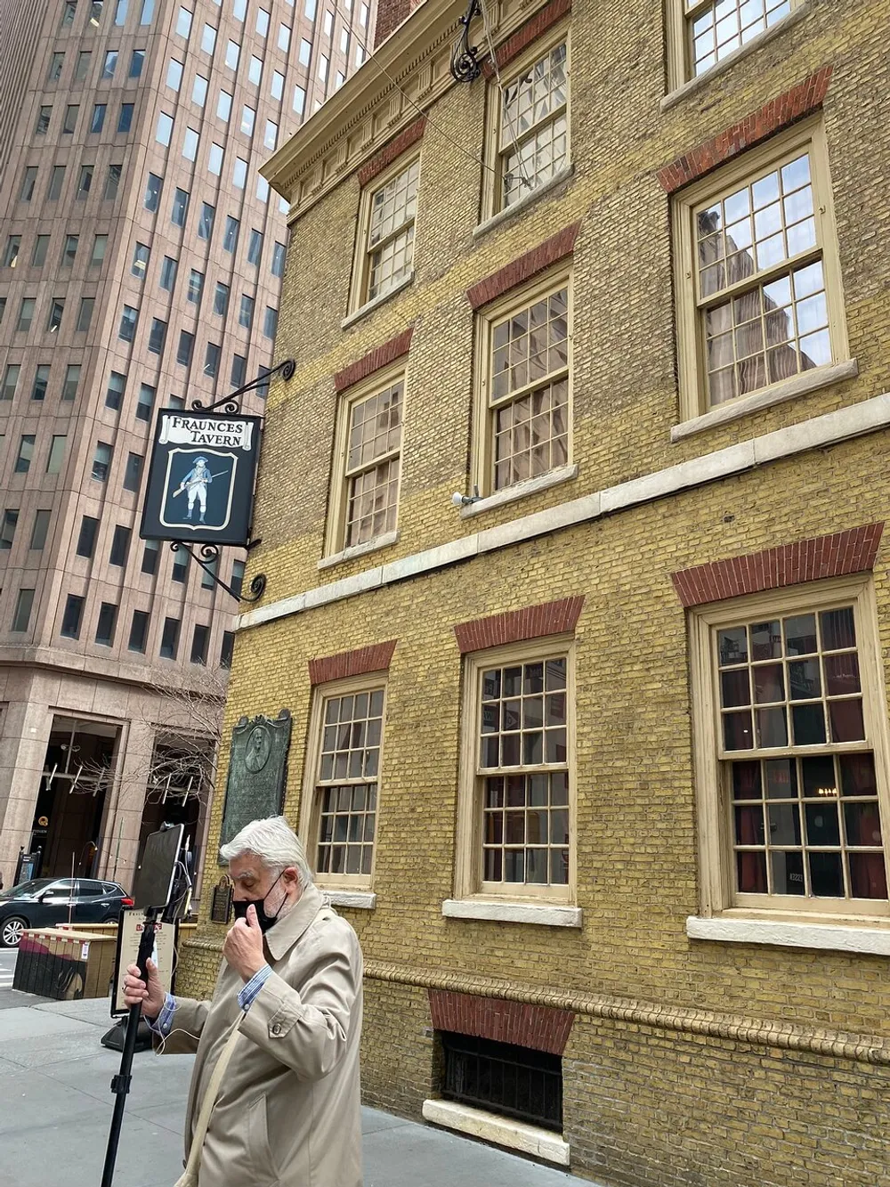 A person is looking at a mobile device in front of Fraunces Tavern with a historical building plaque situated amid contrasting modern architecture