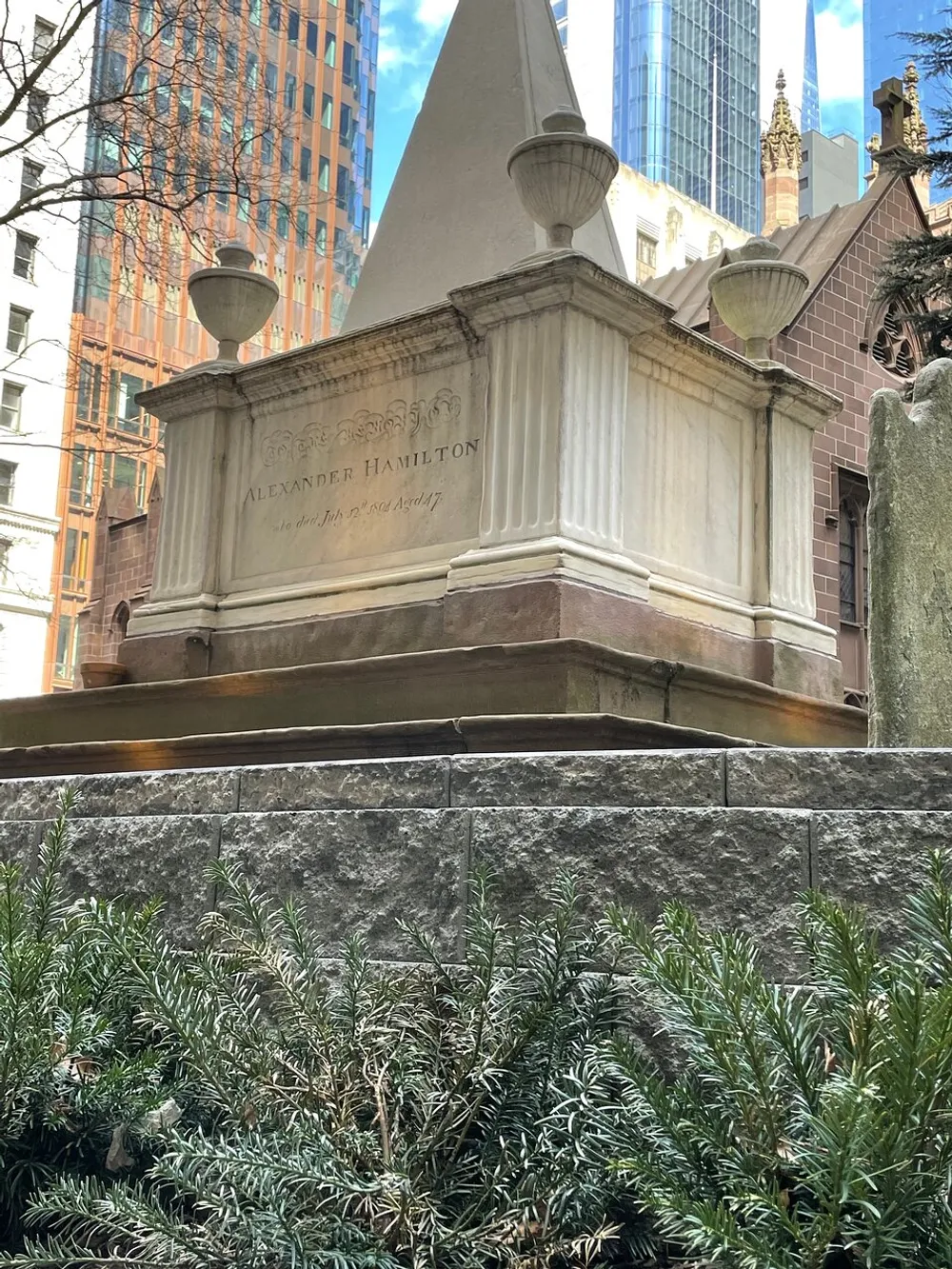 This image features the grave of Alexander Hamilton marked by a neoclassical monument surrounded by greenery with modern skyscrapers in the background highlighting a juxtaposition between historical legacy and contemporary urban environment