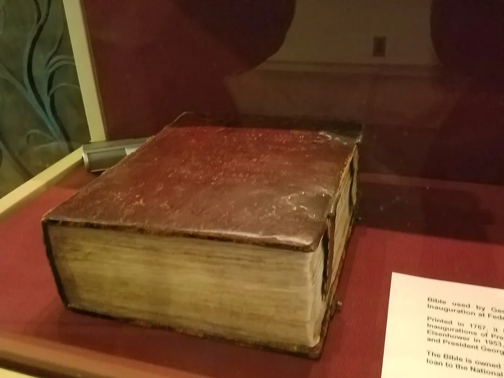 The image shows a large old leather-bound book displayed in a glass case which appears to have historical significance based on its presentation and accompanying information plaque