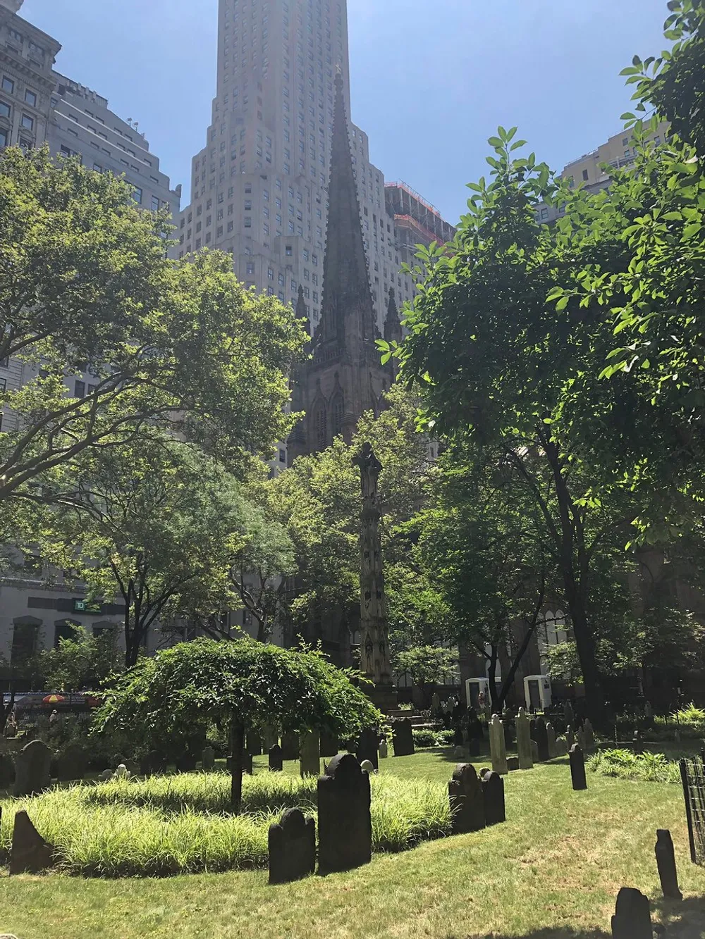 The image shows an urban cemetery with weathered headstones nestled among greenery in the shadow of towering skyscrapers highlighting a contrast between the tranquility of the resting place and the bustling cityscape