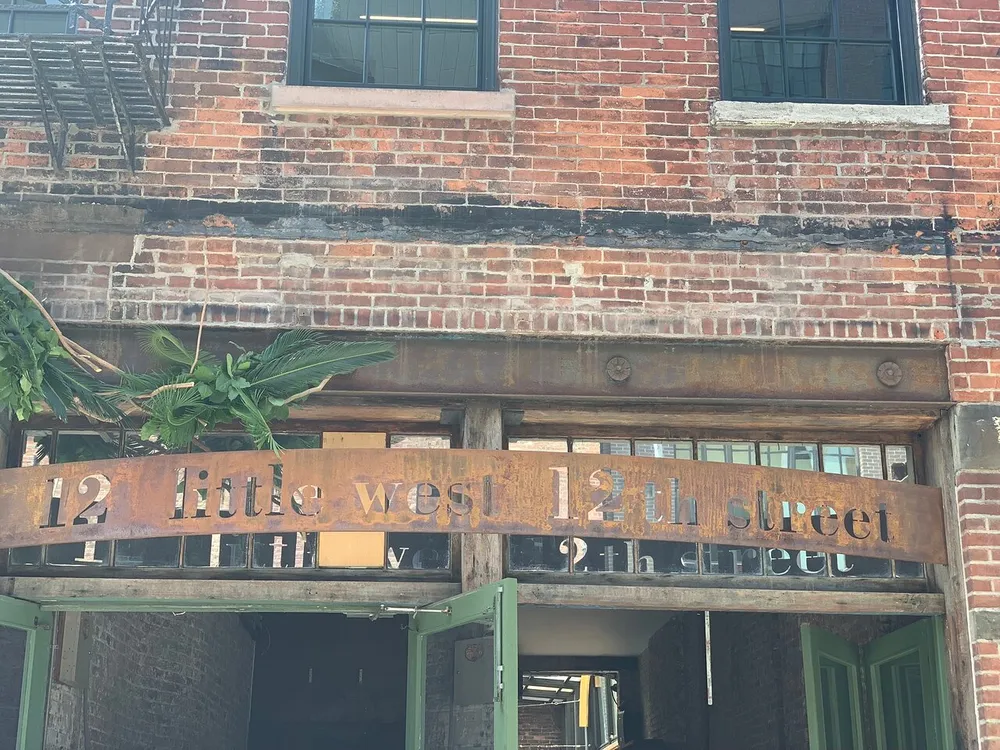 The image shows a rusted metal sign with cut-out letters reading 12 little west 12th street affixed above the entrance of a red brick building with dark green doors and a fire escape