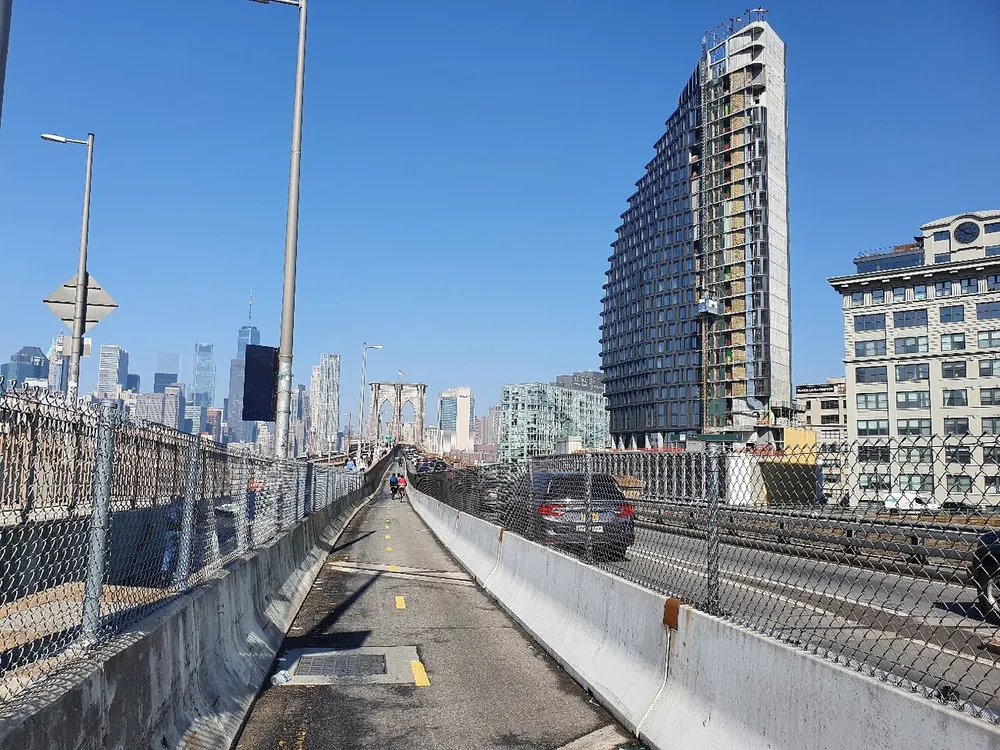 A pedestrian walkway runs alongside a busy road on a bridge with a view of the city skyline and nearby buildings under a clear blue sky