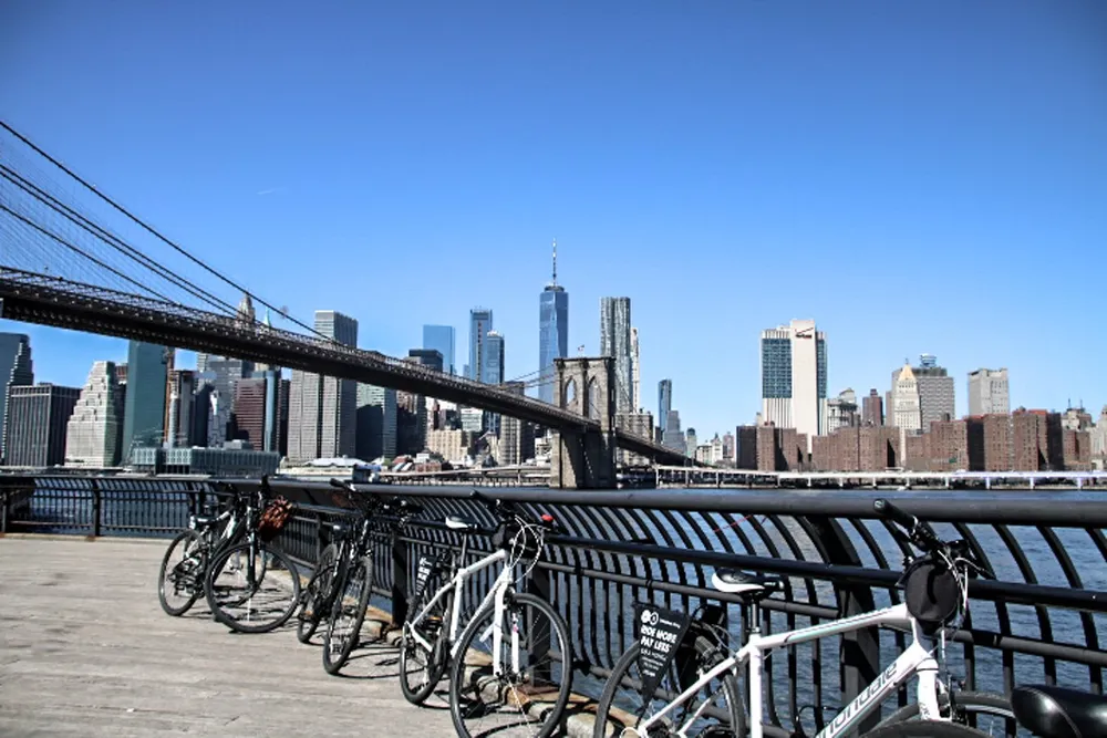 Bicycles are parked along a railing with the Brooklyn Bridge and the New York City skyline in the background on a clear day