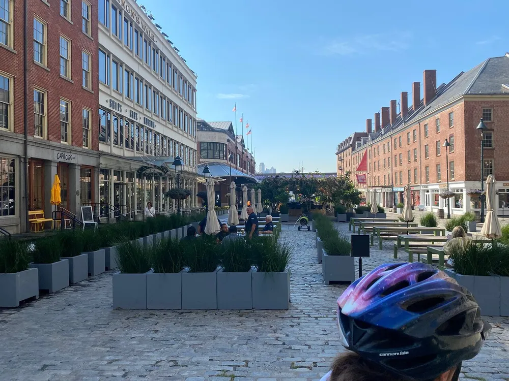 A person wearing a bicycle helmet is looking down a pedestrianized cobblestone street lined with buildings and outdoor seating areas under clear blue skies