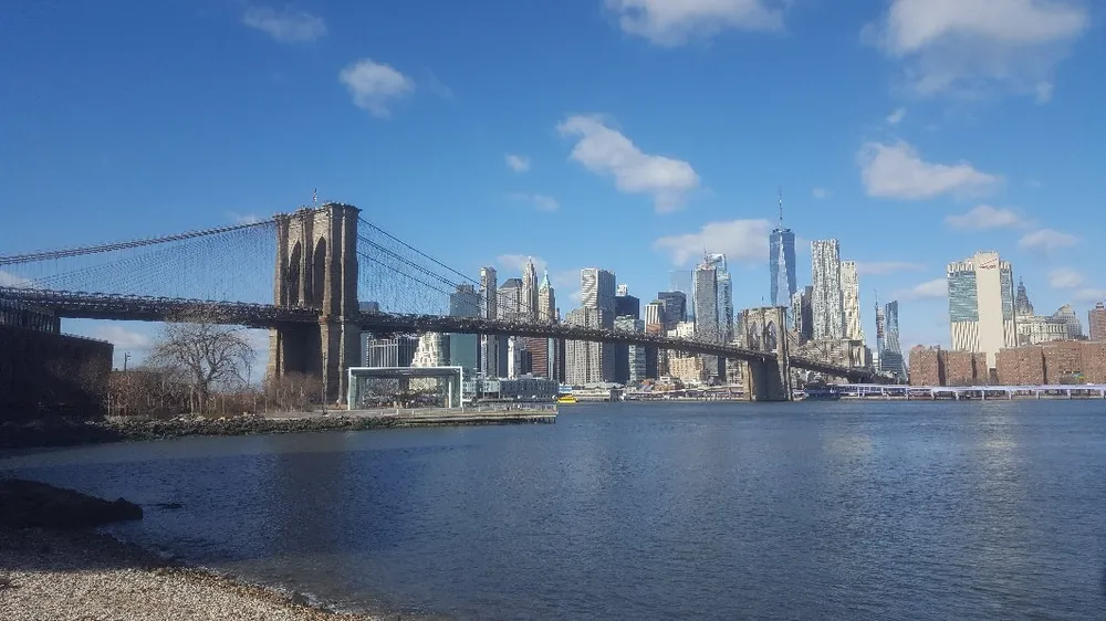 The image shows the Brooklyn Bridge spanning the East River with the Manhattan skyline in the background under a clear blue sky
