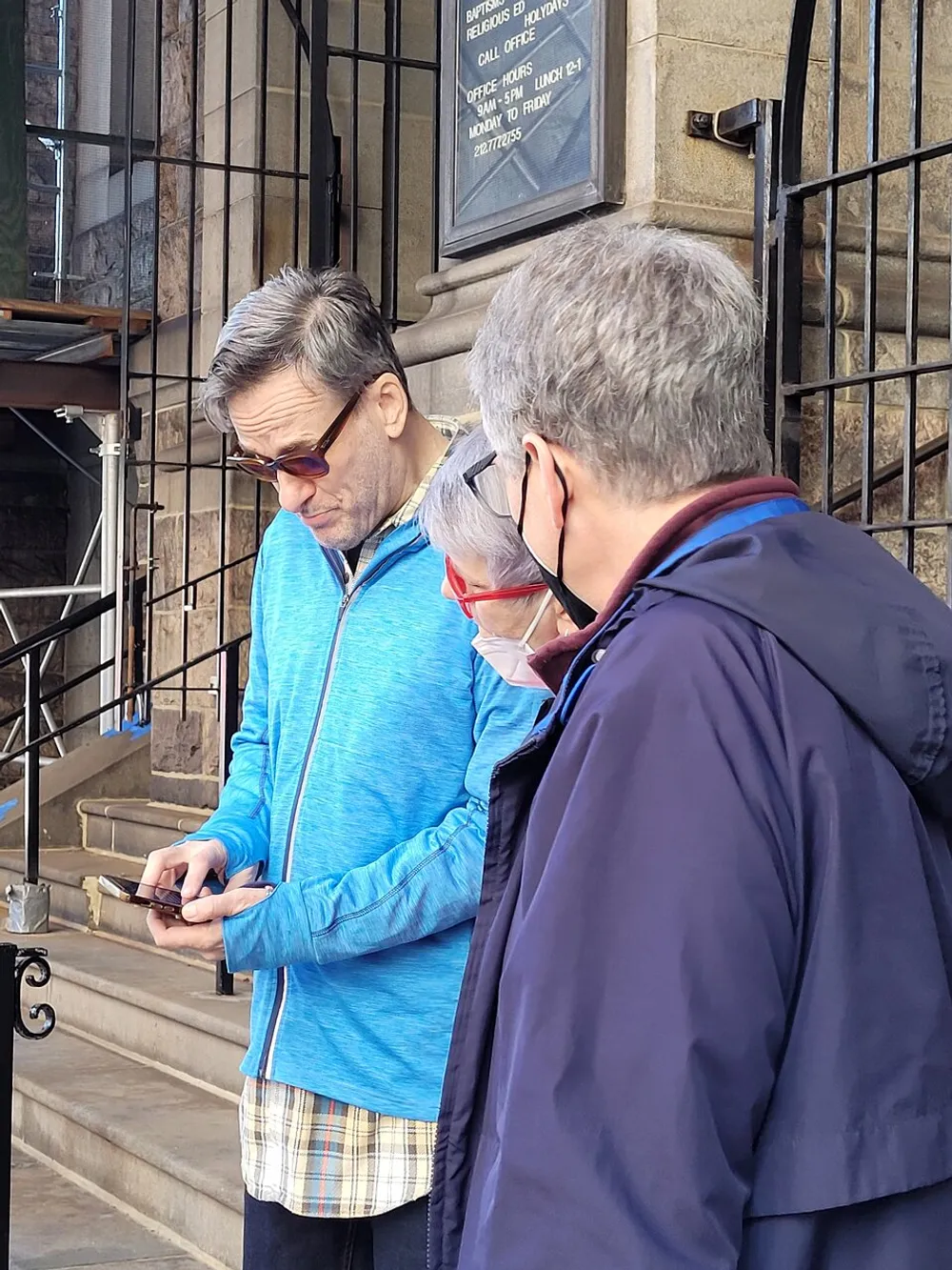 Three people appear engrossed in looking at something on a smartphone outside a building with a sign indicating office hours for religious services