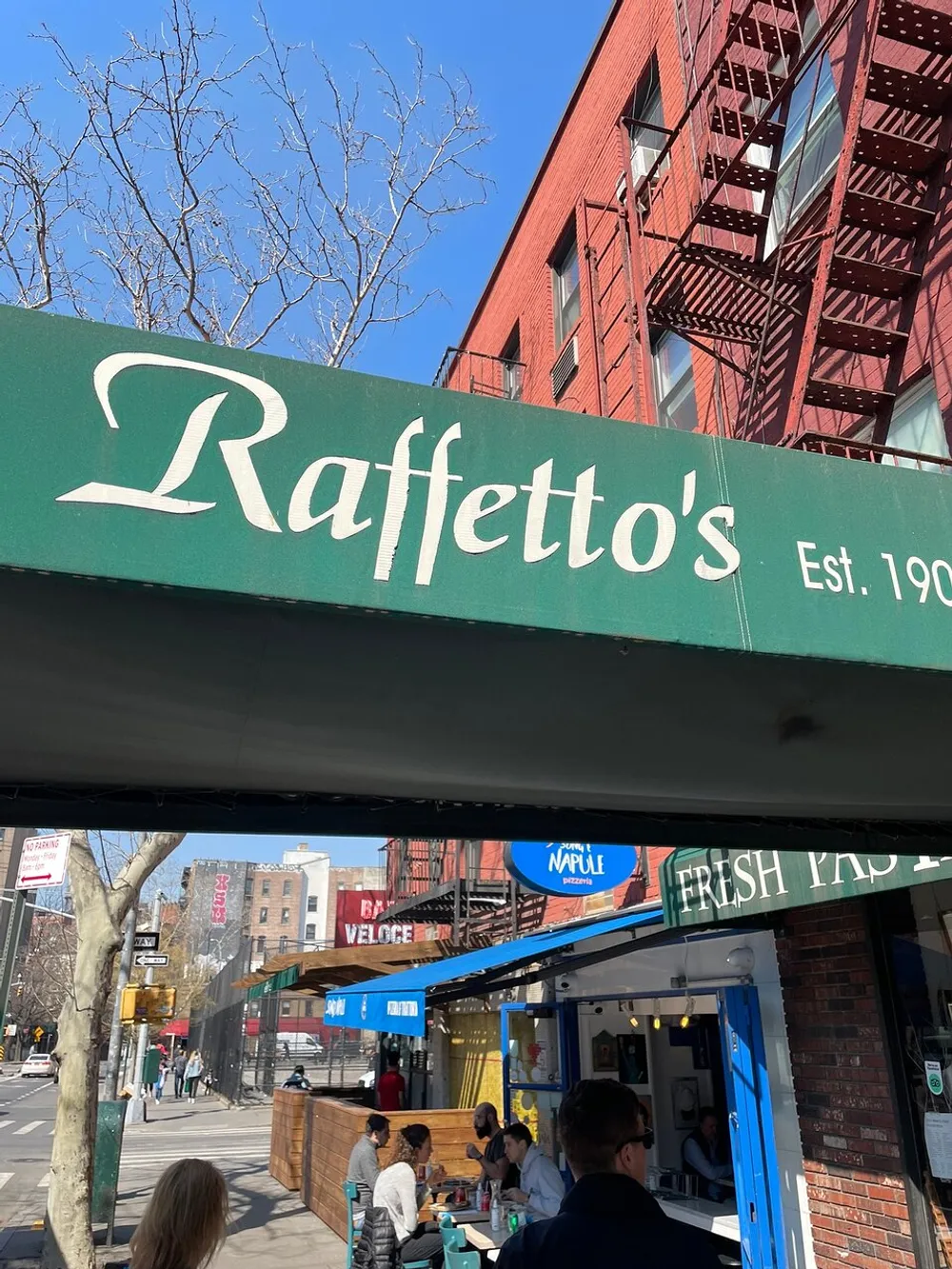The image displays the storefront of Raffettos established in 1906 with people dining at an outdoor seating area under a clear blue sky