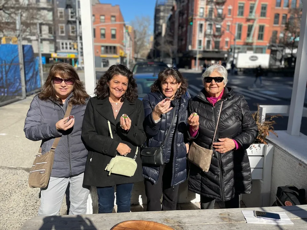 Four women are posing with smiles for a photo on a sunny day on a city street each holding a small object in their hand