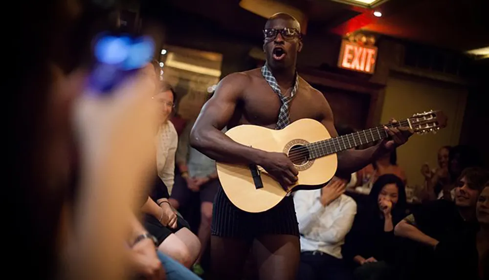 A shirtless man wearing glasses and a tie plays the guitar for an amused audience