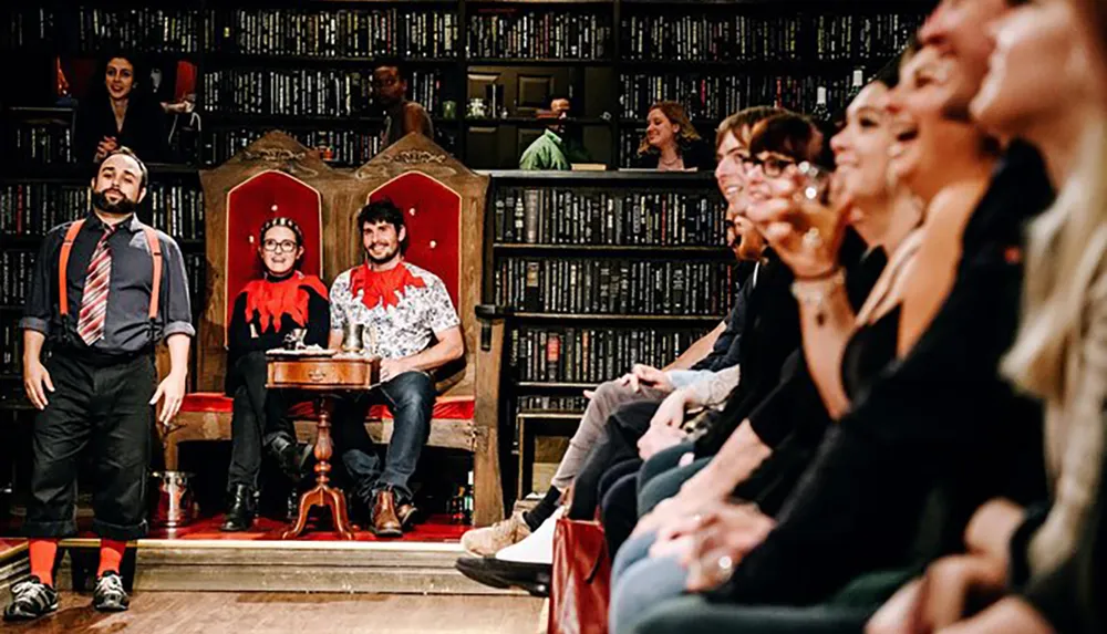 The image shows a lively gathering of people with two individuals sitting on thrones at a table another person standing with a microphone and spectators in the background all set against a wall of bookshelves