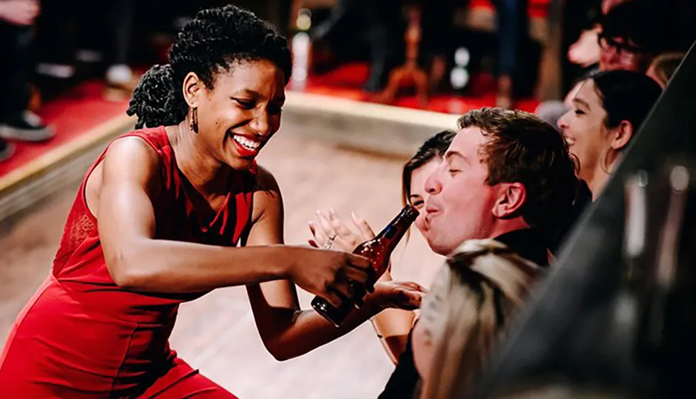 A woman in a red dress is laughing and clinking a beer bottle with a man who is making a funny face as they enjoy a jovial moment at a social gathering