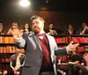 A man in a suit with a bow tie is animatedly addressing an audience in an intimate venue with bookshelves in the background