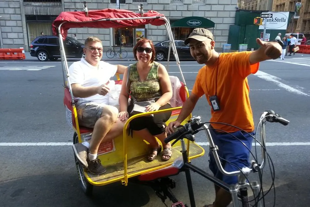A rickshaw driver gives a thumbs-up while two passengers smile at the camera in the back seat of a pedicab on a sunny city street