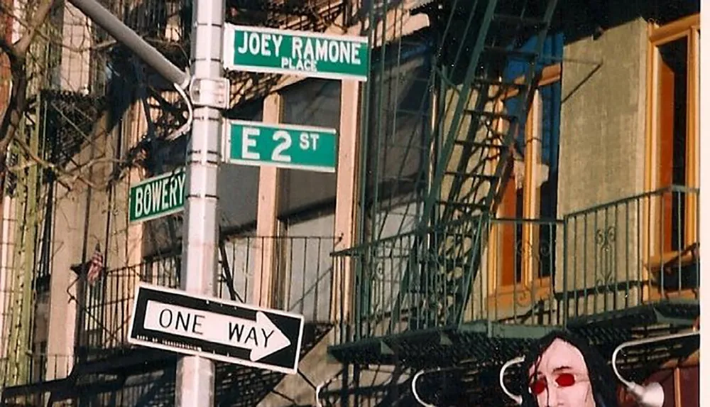 The image shows street signs at an intersection including one named Joey Ramone Place at the corner of Bowery and East 2nd Street with a building and fire escape in the background