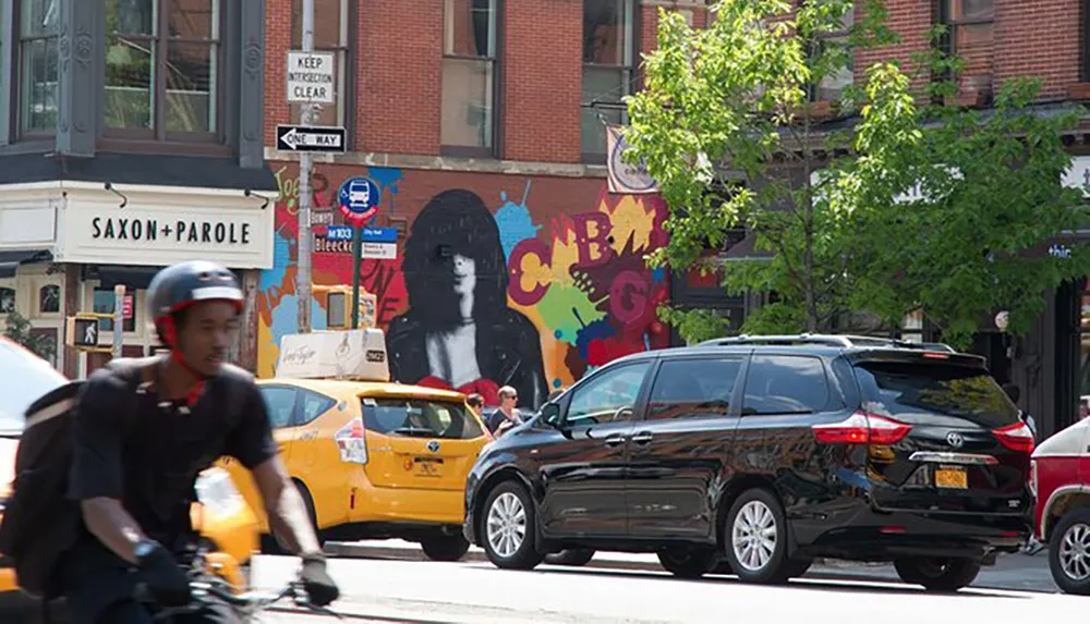 A cyclist in the foreground moves past a busy city street with cars and taxis with a vibrant colorful mural on a building in the background
