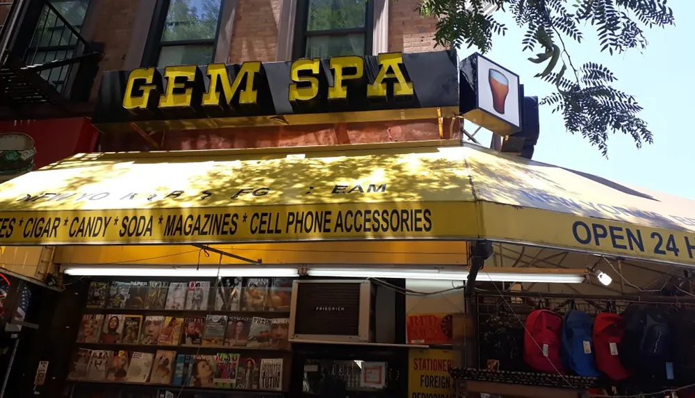 The image shows the exterior of Gem Spa a storefront advertising various items such as cigars candy soda magazines and cell phone accessories and indicating that it is open 24 hours a day
