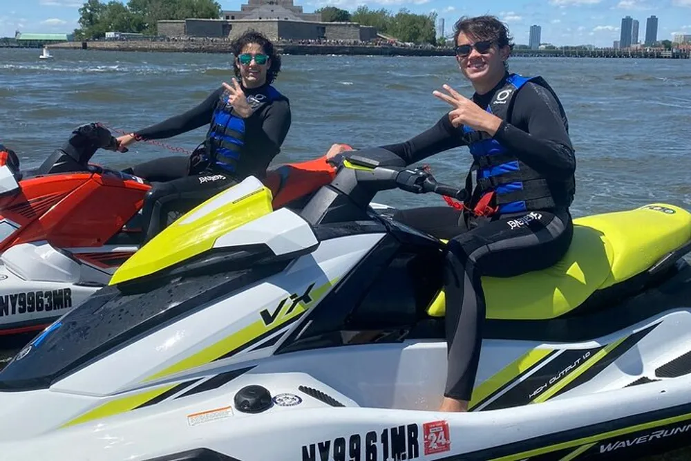 Two individuals in wetsuits are seated on personal watercraft smiling and making peace signs with a waterfront and blue sky in the background