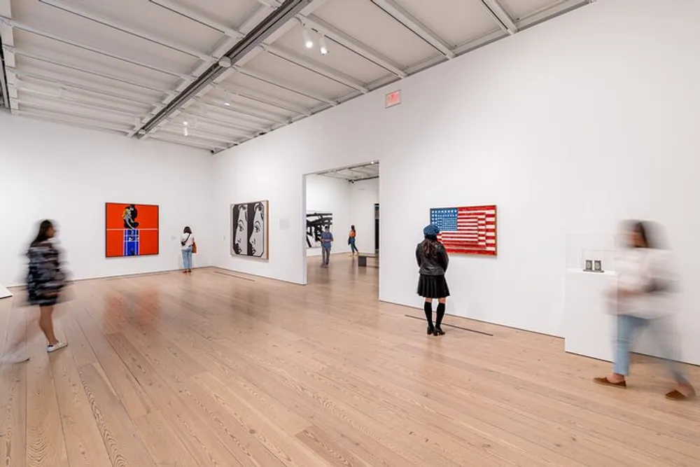 This image depicts visitors observing artwork in a modern gallery space with white walls wooden floors and various pieces of art including an American flag-themed piece