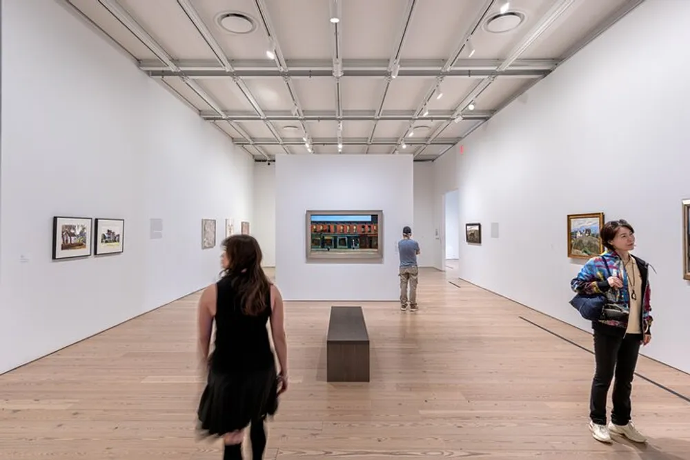 Visitors are viewing art exhibits in a modern gallery with white walls wooden floors and framed paintings hung at eye level
