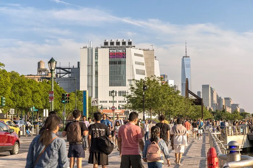 People are walking along a busy waterfront promenade with the Whitney Museum and the New York City skyline in the background