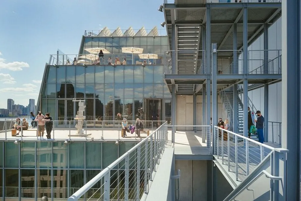 This image shows people enjoying a sunny day on a modern rooftop terrace with glass structures and a view of a city skyline