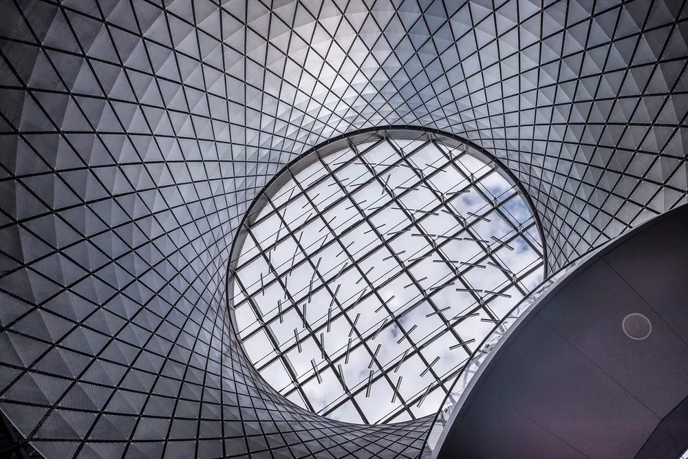 This image features a geometric ceiling structure with a large circular oculus revealing a patterned network of beams against a cloudy sky