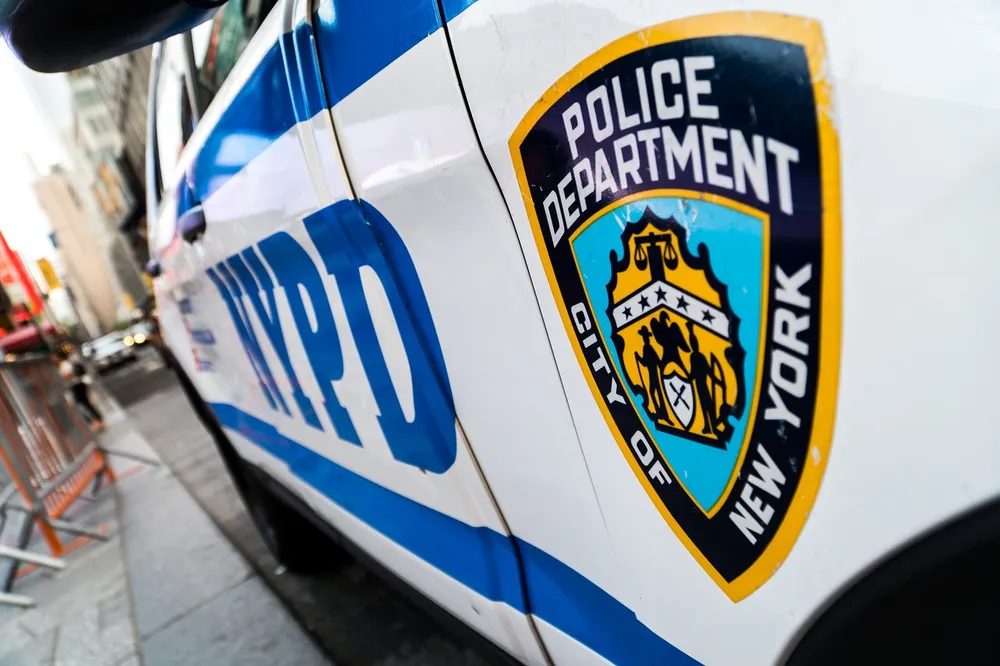The image shows a close-up of the emblem on a New York Police Department NYPD vehicle with urban surroundings intentionally blurred in the background