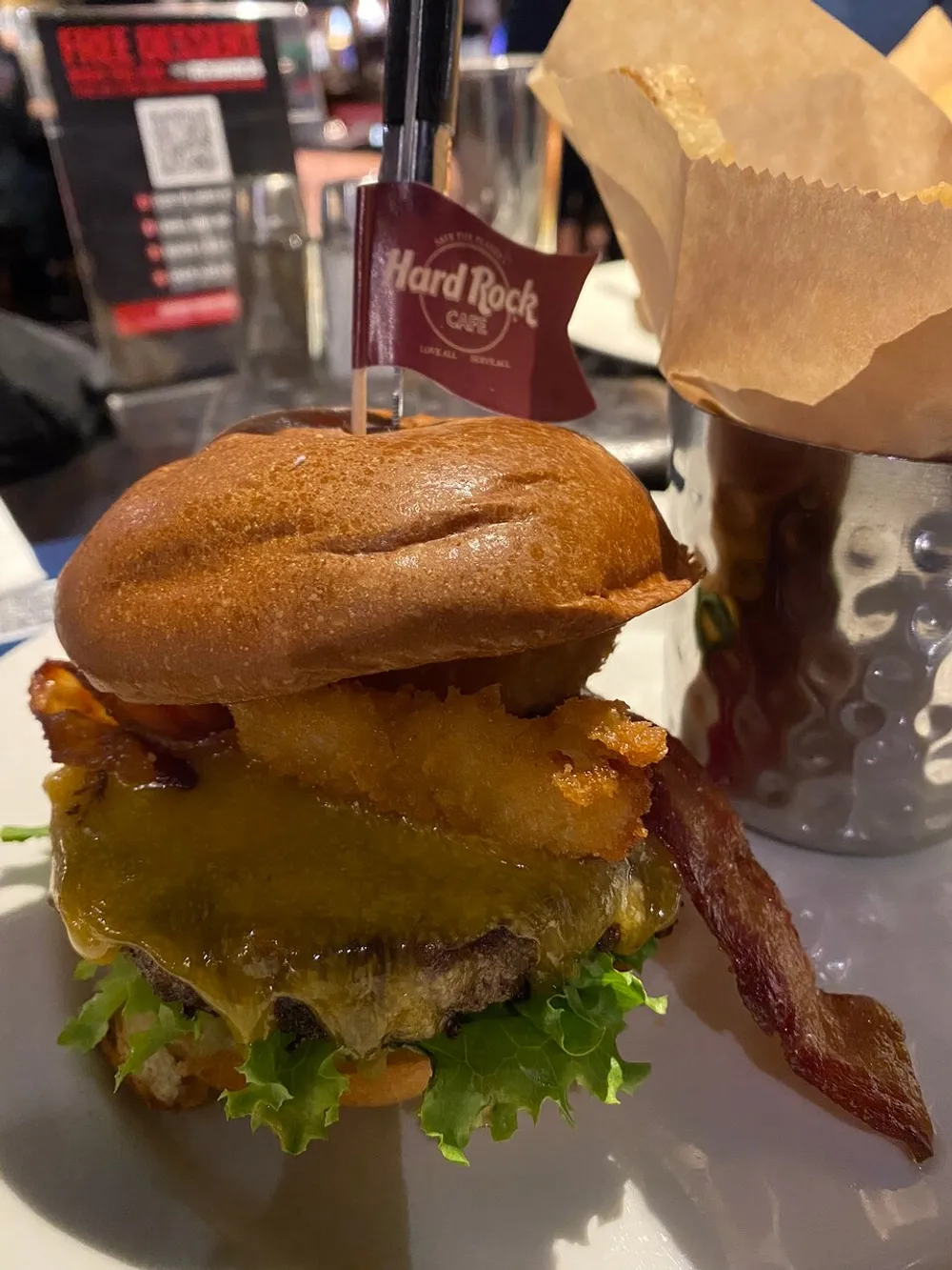 The image shows a gourmet burger with cheese bacon lettuce and a fried onion ring skewered with a branded toothpick from Hard Rock Caf served alongside a metal cup of French fries