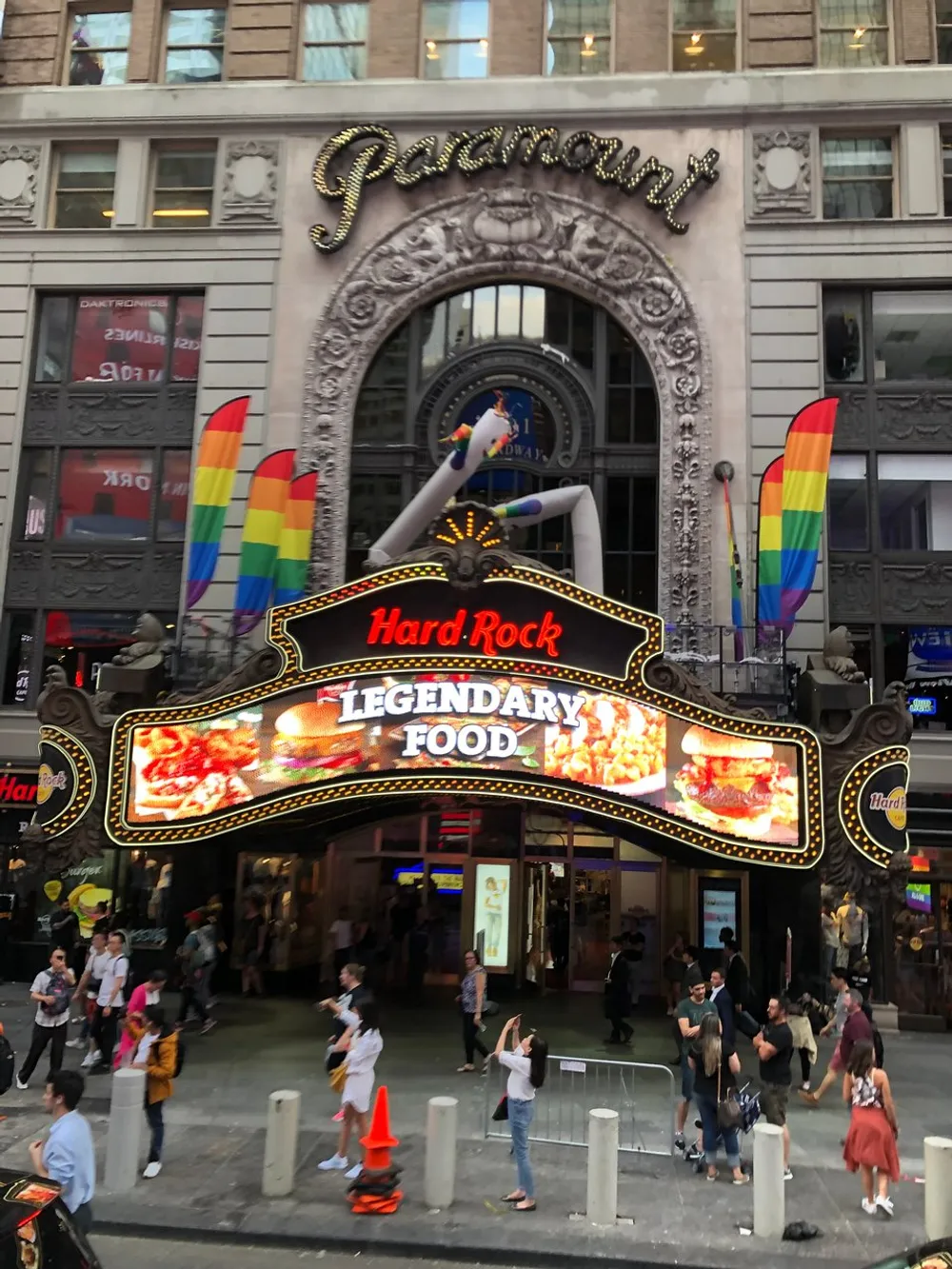 The image shows a busy street scene in front of the Hard Rock Cafe adorned with rainbow pride flags under a vintage-looking Paramount sign