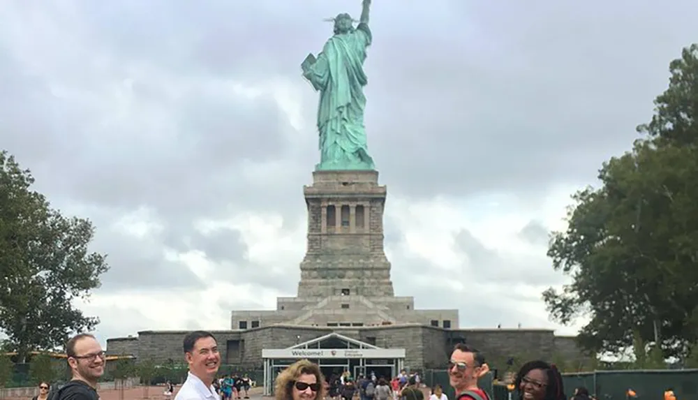 A group of visitors is smiling and posing for a photo in front of the Statue of Liberty on a cloudy day