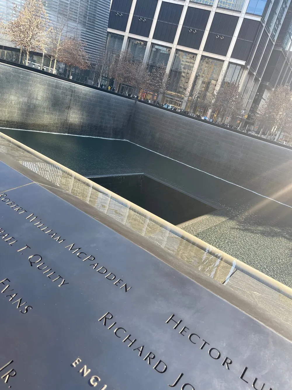 The image shows a reflective pool with water cascading down its sides surrounded by bronze panels inscribed with names creating a solemn and contemplative atmosphere