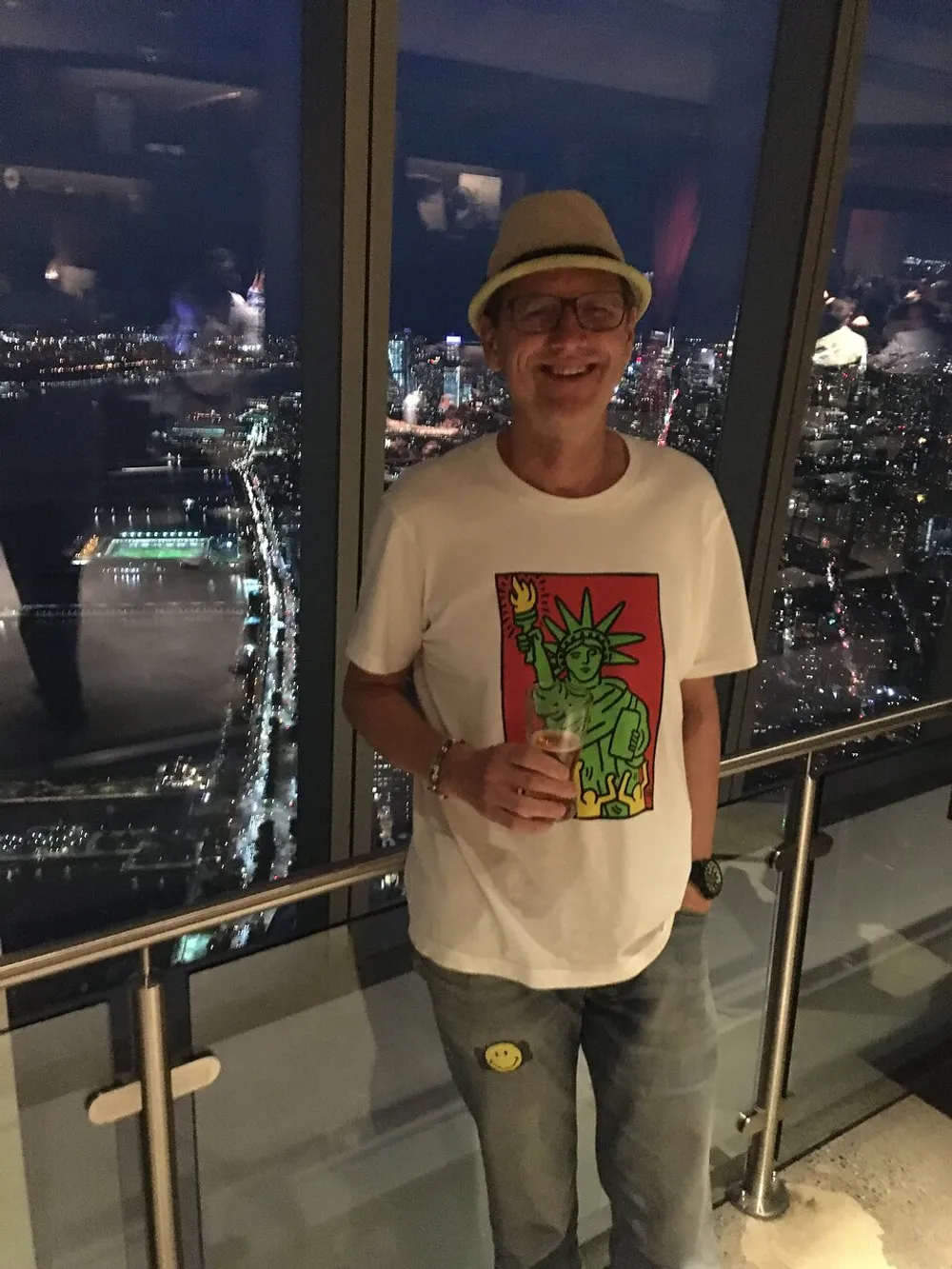 A smiling person wearing a hat and a T-shirt with a colorful graphic stands indoors with a night cityscape visible through large windows in the background