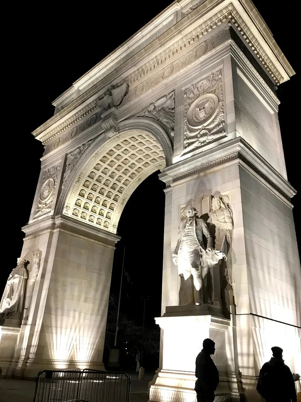 The image shows the Washington Square Arch illuminated at night with sculptures adorning its pillars and people standing in the foreground observing it