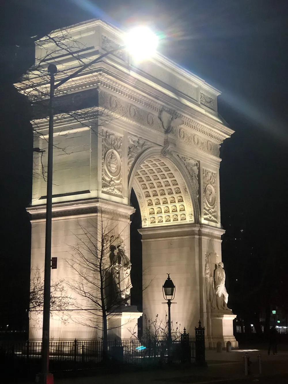 The photo shows a beautifully illuminated arch monument at night with intricate sculptures and a bright street lamp casting a glow on its facade