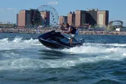 A person is riding a jet ski on the water with a crowded beach and amusement park rides visible in the background.