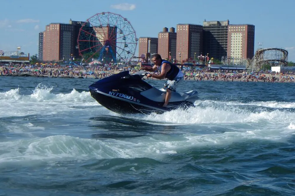 A person is riding a jet ski on the water with a crowded beach and amusement park rides visible in the background